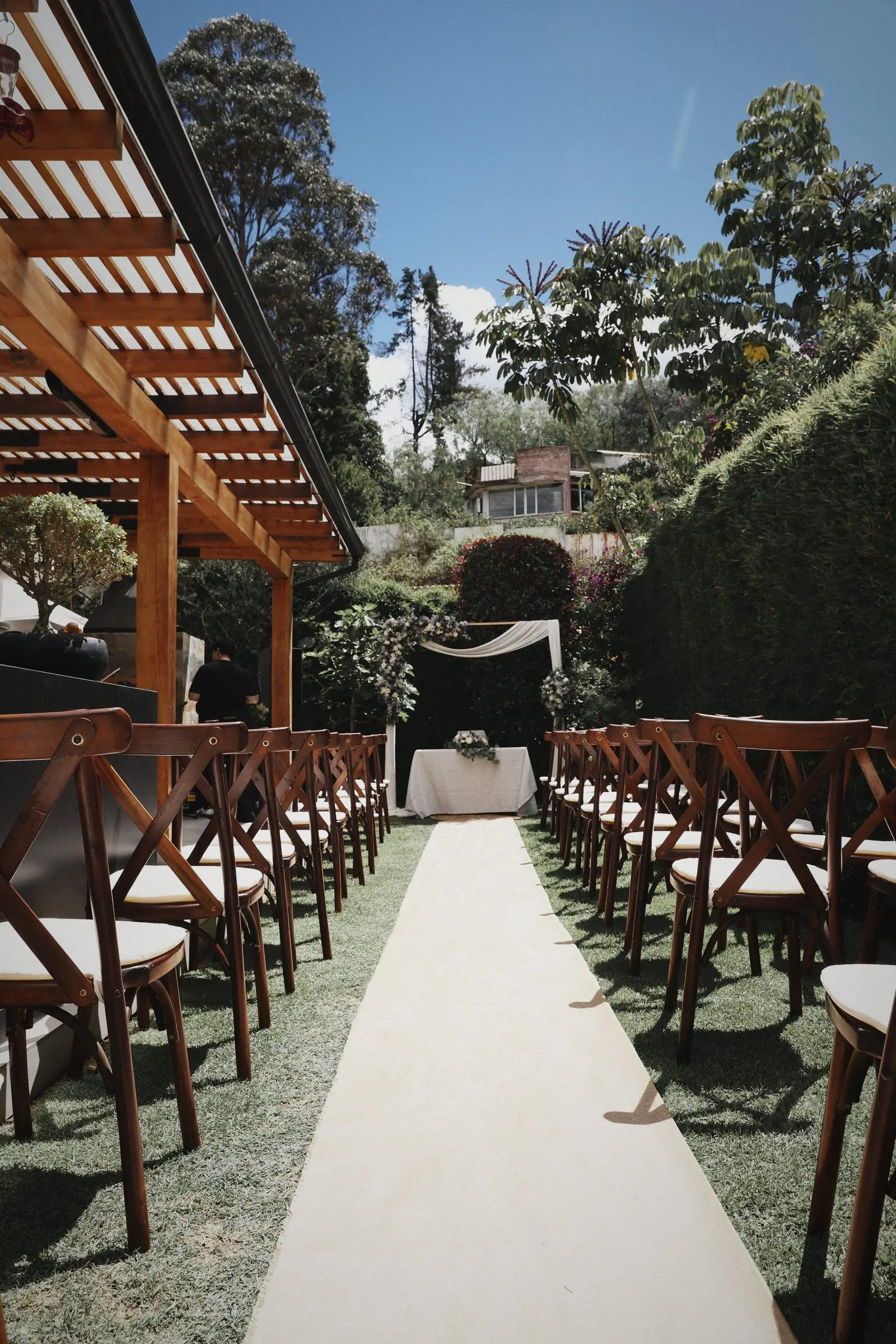 A long aisle of wooden chairs under a timber pergola leading into a garden.